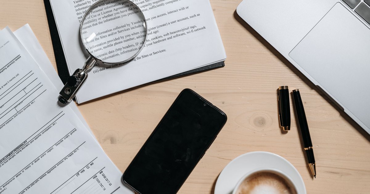 Vista cenital de una mesa de oficina con documentos, una lupa, un portátil y un café