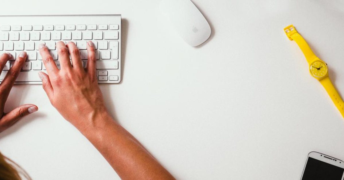Manos escribiendo en un teclado blanco junto a un lápiz amarillo