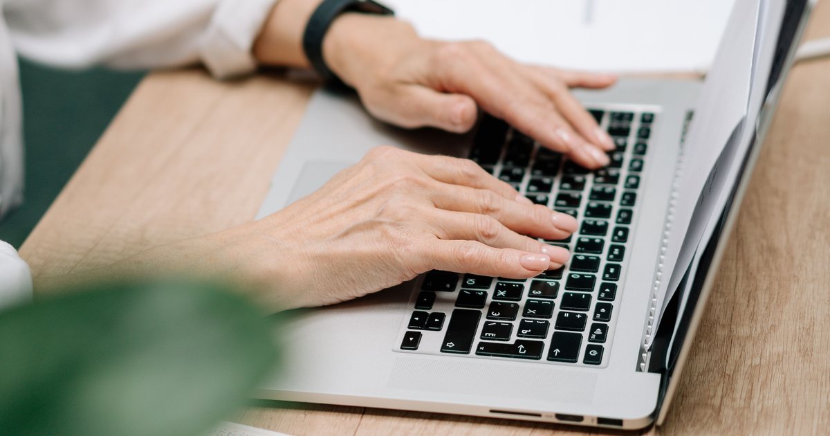 Manos escribiendo en el teclado de un portátil junto a documentos sobre una mesa de madera